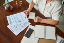 Close up of old lady with pen in her hand sitting at the table with individual income tax return form, cellphone, answer sheet and spiral notebook